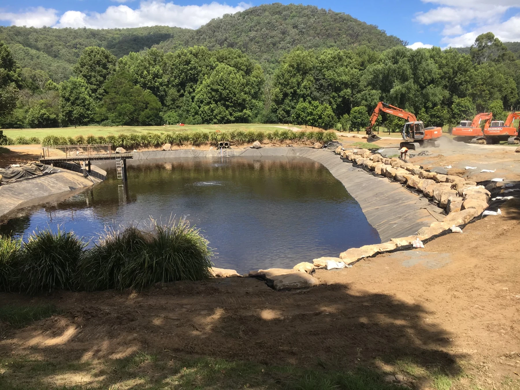 Excavation machinery beside a rural dam with stone edging